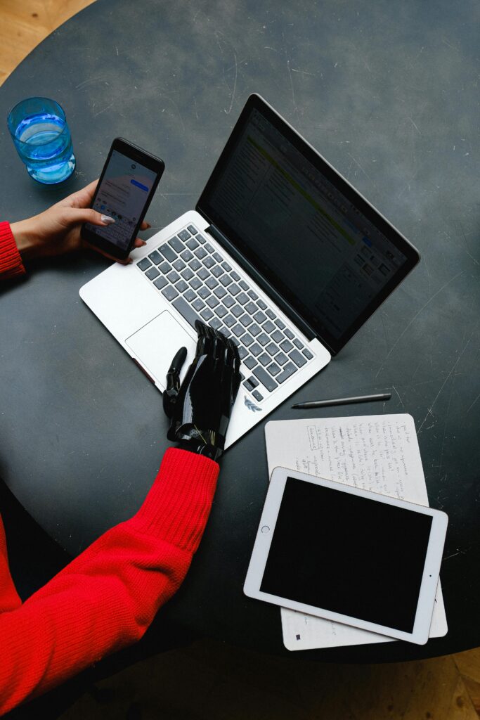 A prosthetic hand uses a laptop on a workspace with gadgets and notes around.